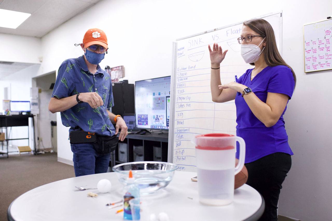 Young man wearing orange Clemson baseball cap stands with Adult Enrichment Staff member behind table with a clear medium sized bowl filled with water during an activity with high5 day program for adults with disabilities