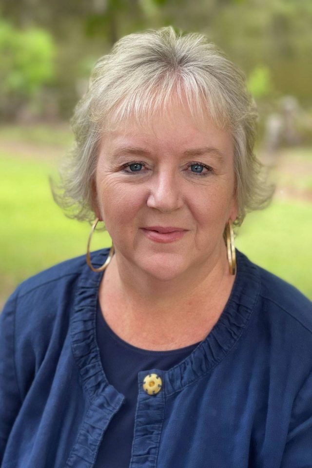 Chief Executive Officer Samantha Kriegshauser smiling into camera with white bobbed hair wearing gold hoop earrings and blue top
