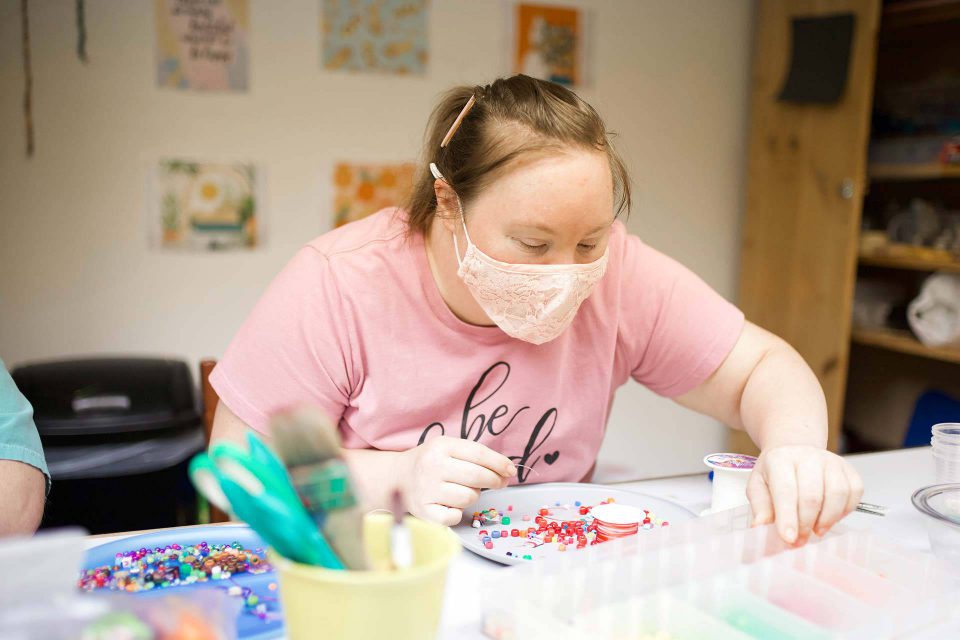 Young woman wearing pink shirt and pale pink lace facemask searches through beads for a bracelet she is constructing during day programs for adults with disabilities