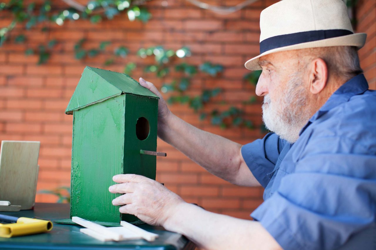 Elderly man in white fedora and blue shirt puts together a wooden birdhouse that has been painted green