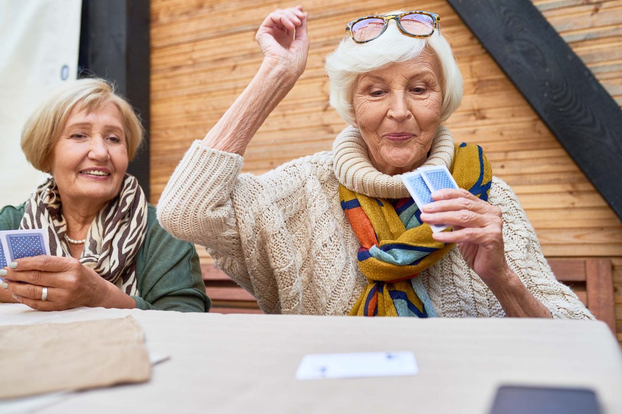 Elderly woman with smart white bob hairstyle and glasses atop her head, wearing a straw colored sweater and a bright orange, ochre, and blue multi-colored scarf looks pleased as she places a card down during a card game.