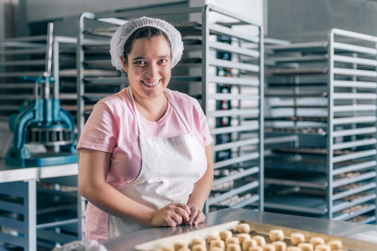 Woman in hair net and apron smiling in front of baking racks