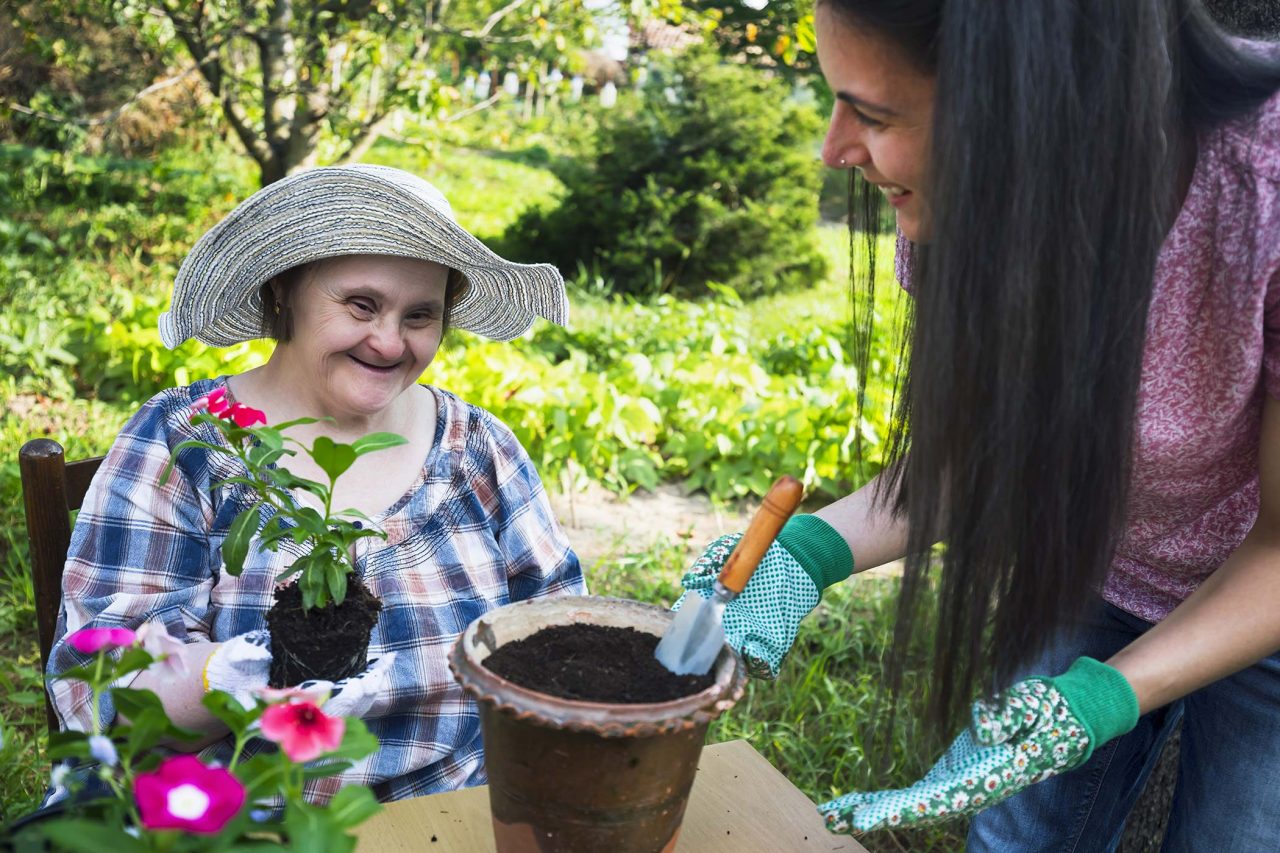 An older woman in a woven sun hat adding a flower to a pot full of soil as another young woman smiles and assists