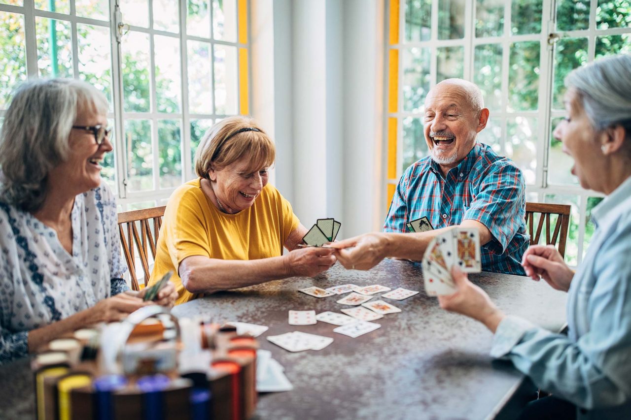 Group of chuckling older individudals plays cards around a table in a brightly lit room