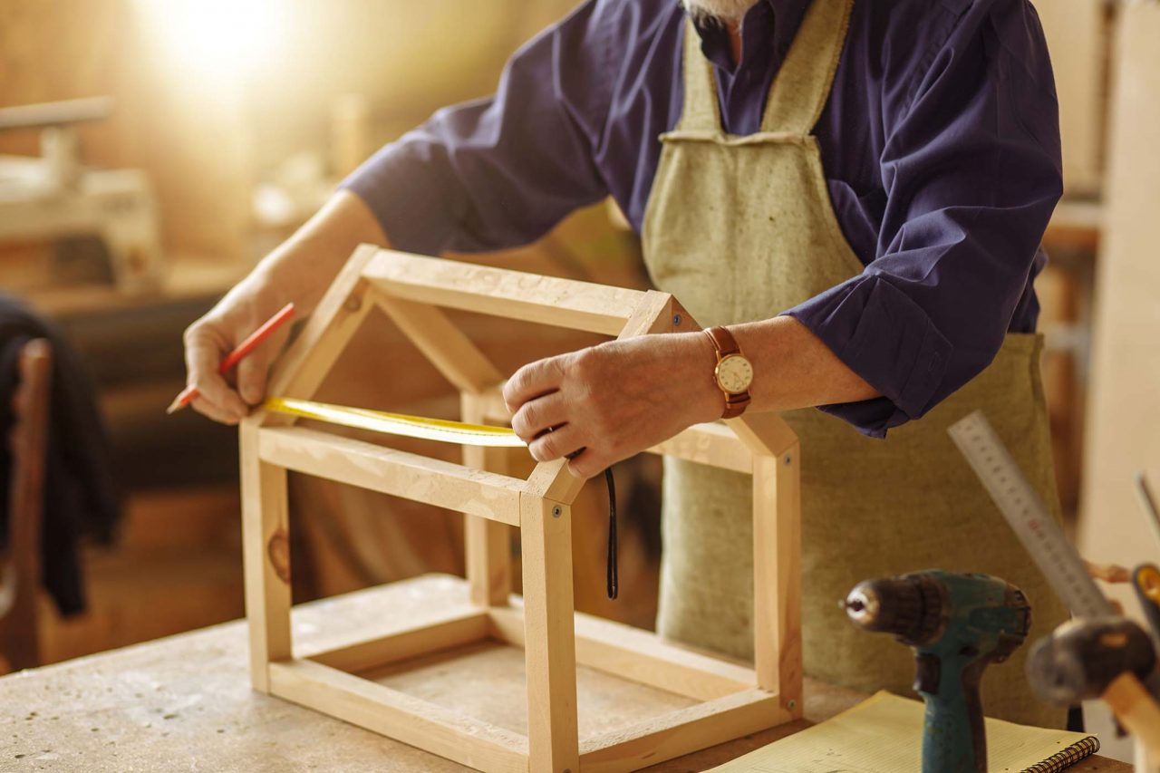 Pair of hands measuring a portion of the wooden frame of a birdhouse in a wood workshop