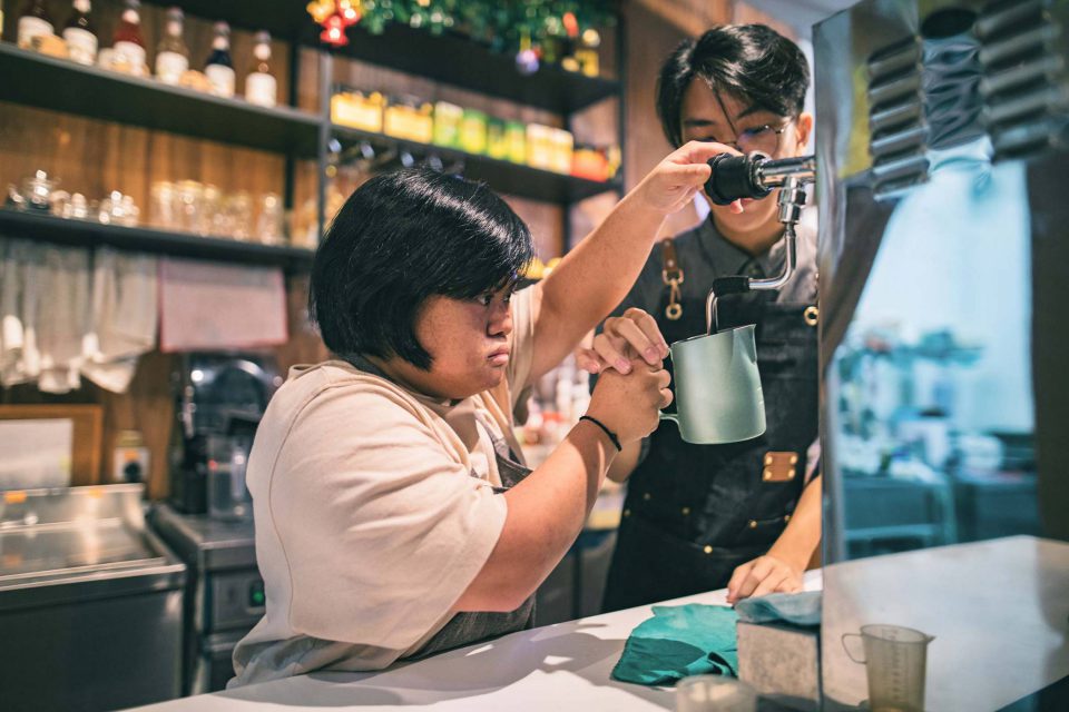 Young woman with cropped shiny dark hair wearing light pink top acting as a barista uses a milk frother assisted by another young male barista at job training for disabled adults