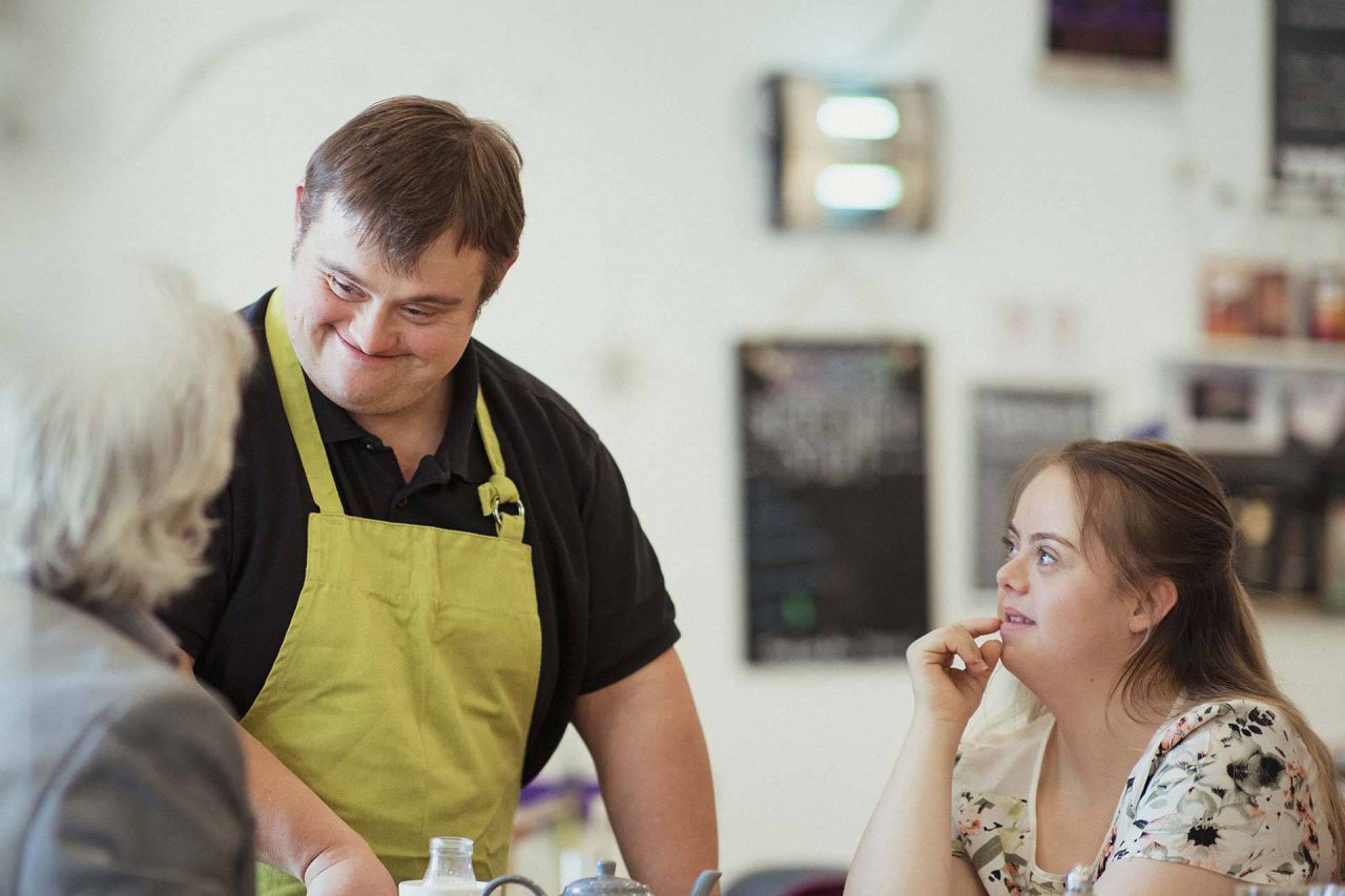 Brown haired waiter at job training for disabled adults wearing black shirt and chartreuse apron smiles at two patrons