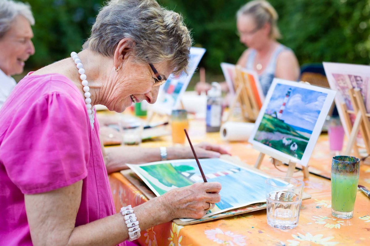 Elderly woman with short gray bob, wearing fuchsia blouse, thick white beaded necklace, and glasses, painting a watercolor of a lighthouse at adult day care with others.¬¬¬¬