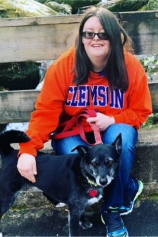 Smiling young woman with dark hair wearing an orange Clemson sweatshirt has her arm around a small black dog