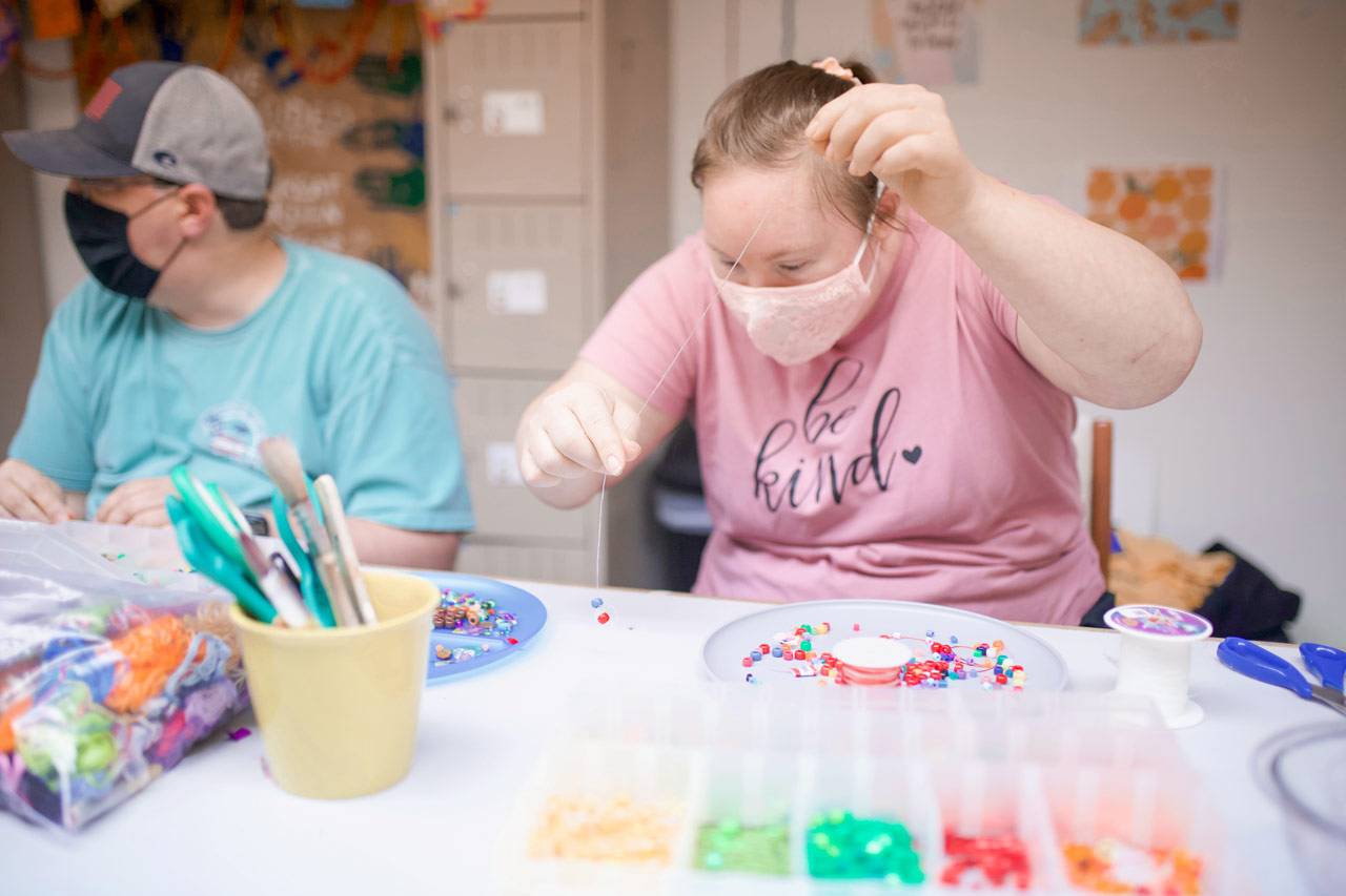 Young woman wearing pink shirt and pale pink lace facemask placing beads on a string during day programs for adults with disabilities. Another program member, a male in a blue shirt and hat, is sitting to her right