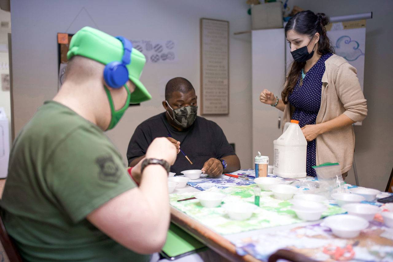 Members of High5 programs for adults with disabilities sit at table covered with several white Styrofoam bowls completing an activity while a female volunteer watches