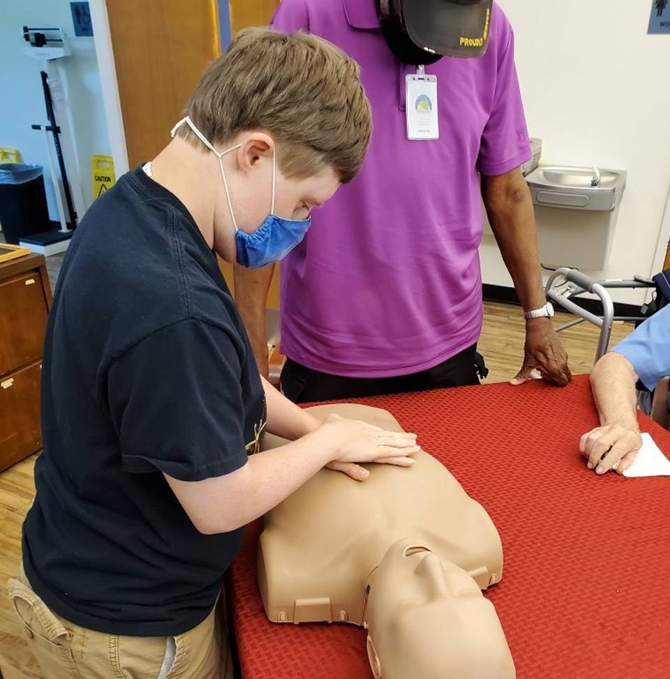 Young male thrive adult daycare member with hands placed to give CPR compressions to plastic dummy
