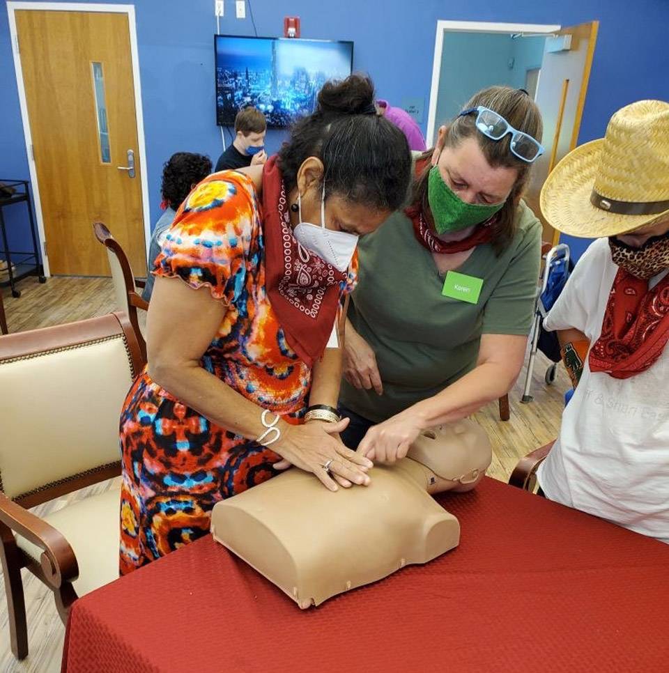 Older female thrive adult daycare member with hands placed to give CPR compressions to plastic dummy assisted by female volunteer as an individual in a straw cowboy hat observes