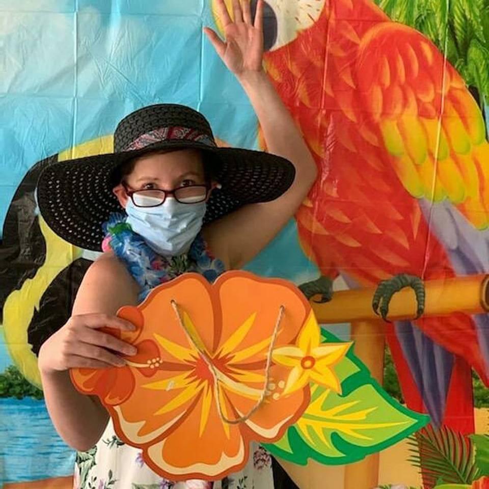Female thrive adult daycare member wearing leis holds colorful cardboard Hawaiian Hibiscus in front of tropical plastic background